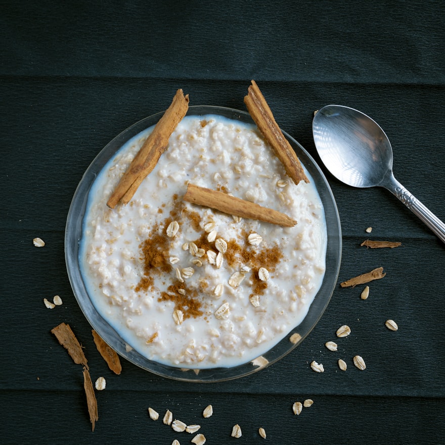 Bowl of creamy oatmeal topped with cinnamon sticks and ground cinnamon, scattered oats and spoon beside it on dark wooden surface.