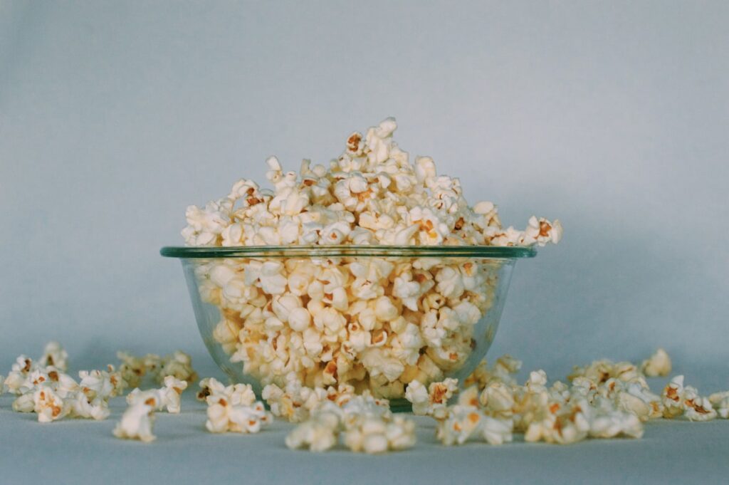 Glass bowl overflowing with buttery popcorn, kernels scattered on pale blue surface.