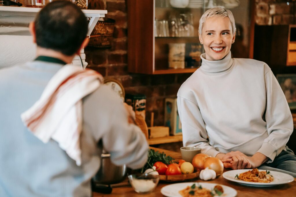 Woman with short white hair in a gray turtleneck smiling at someone across a table with fresh vegetables and pasta.