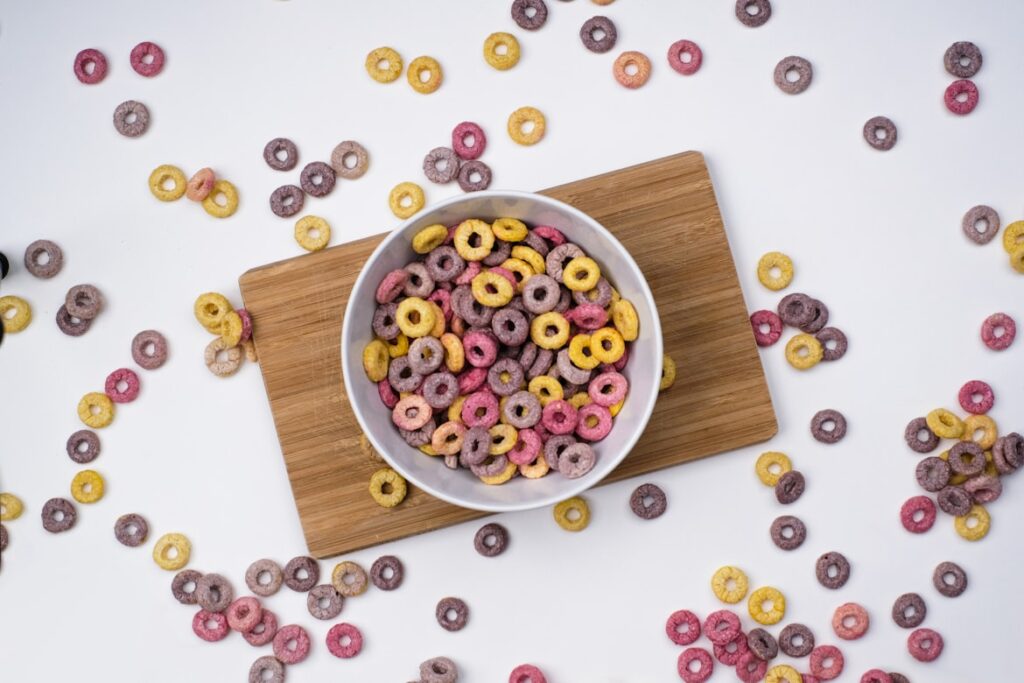Colorful ring-shaped cereal in pink, yellow, and purple scattered across white surface and filling a white bowl on wooden board.