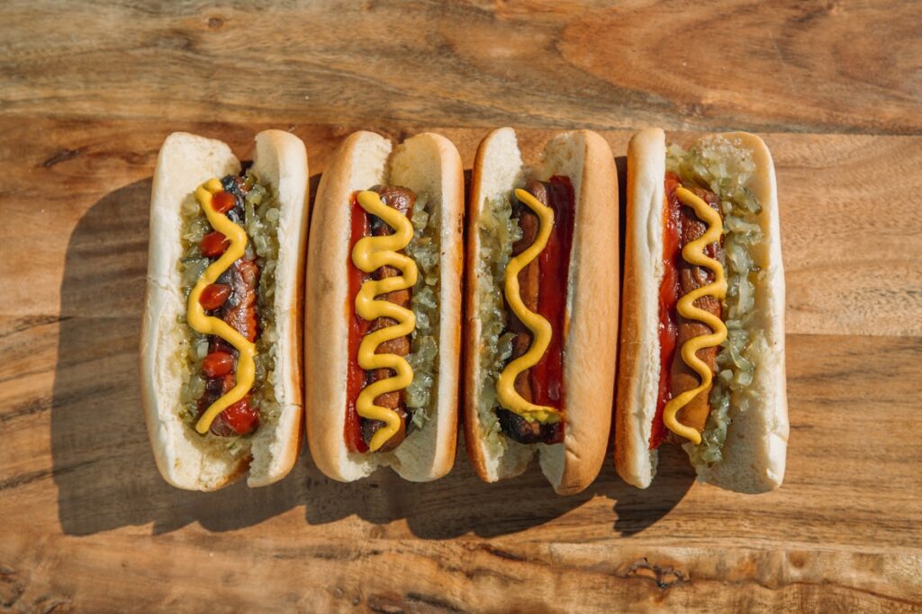 Five hot dogs in buns topped with yellow mustard and green relish, lined up on wooden cutting board.