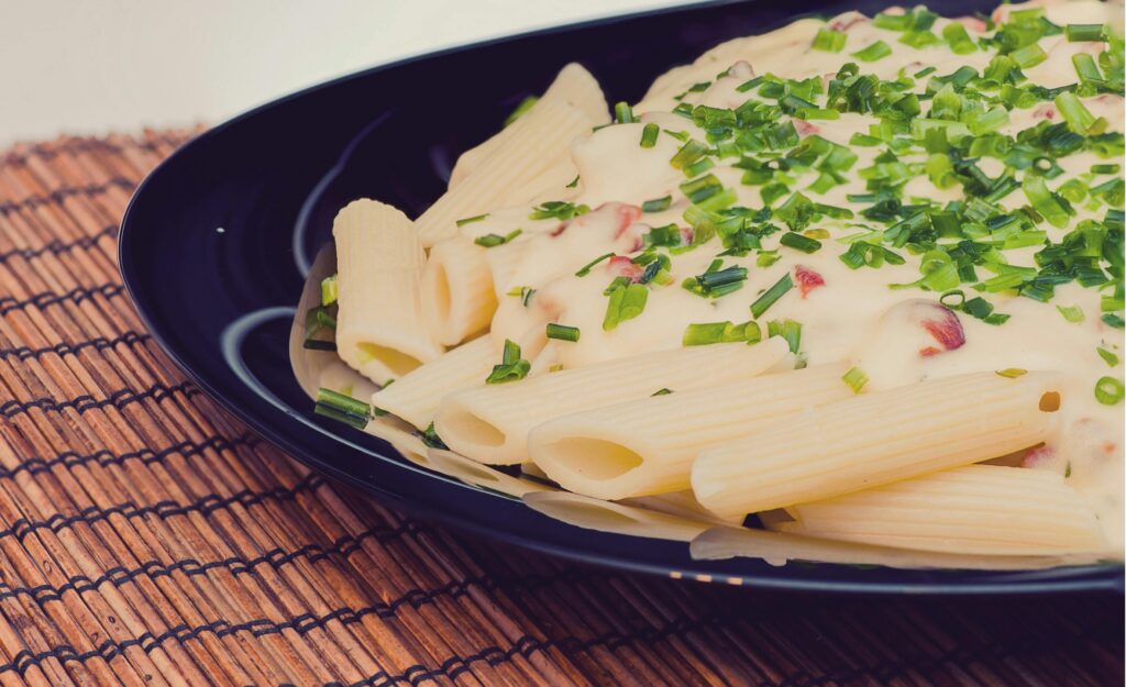 Penne pasta in creamy white sauce topped with chopped fresh chives on dark blue plate.