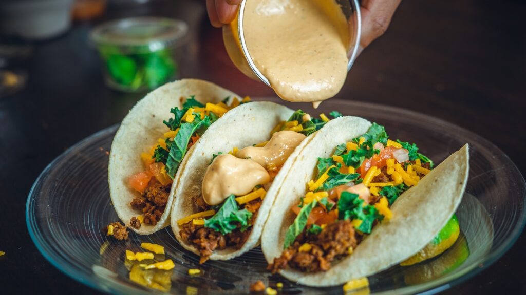 Three soft tacos filled with seasoned ground meat, shredded cheese, and kale, with creamy orange sauce being drizzled from small container.