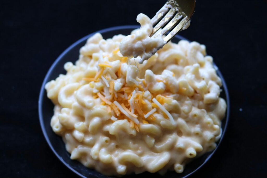 Creamy macaroni and cheese topped with shredded cheddar, fork lifting a bite from dark blue bowl against black background.