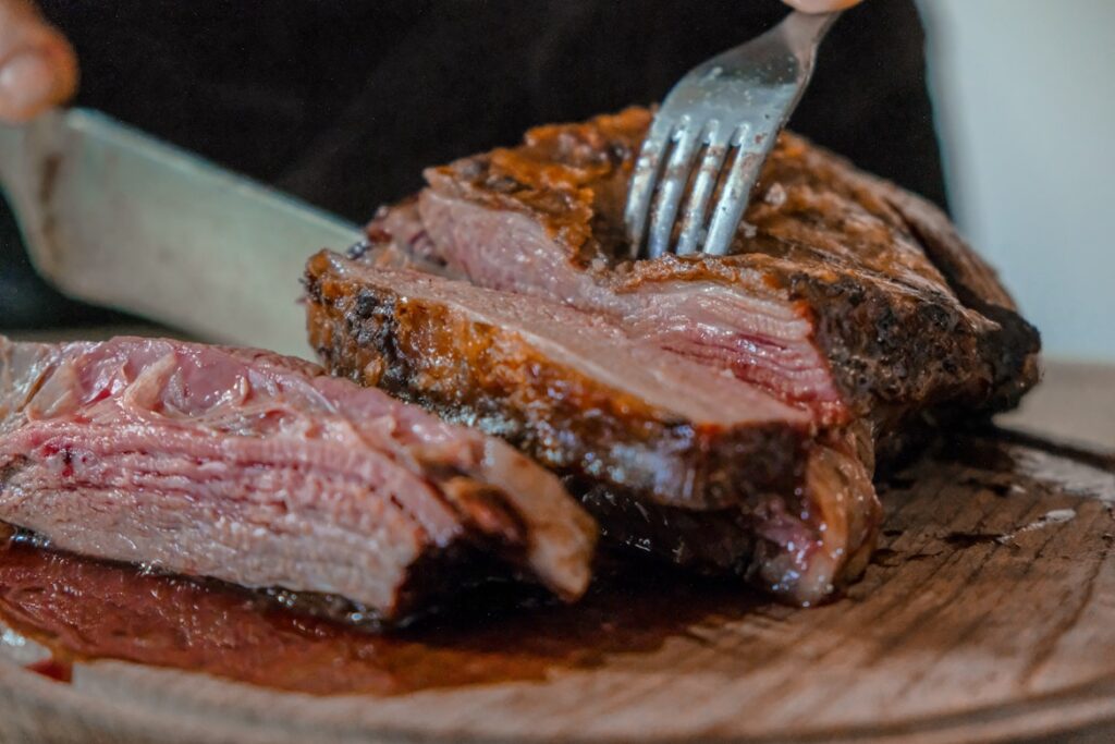 Medium-rare steak being sliced on wooden board, showing pink interior and caramelized crust, fork and knife in frame.