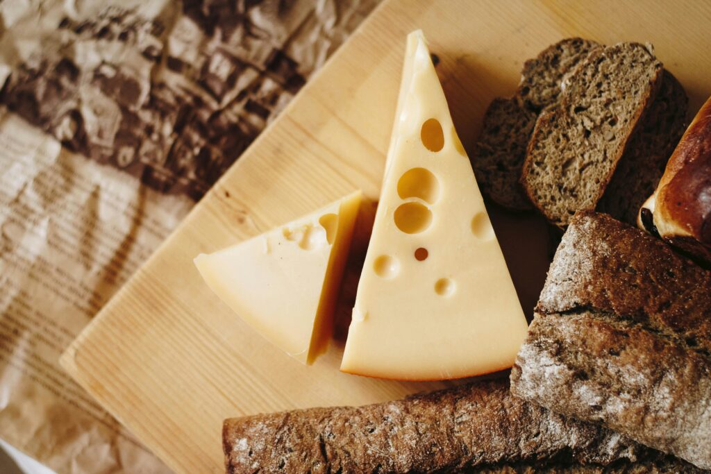 Two wedges of Swiss cheese with characteristic holes beside slices of dark rye bread on wooden cutting board.