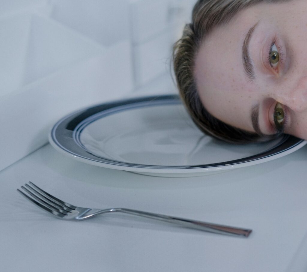 Person resting their head at table level, gazing at empty silver plate with fork beside it on white surface.