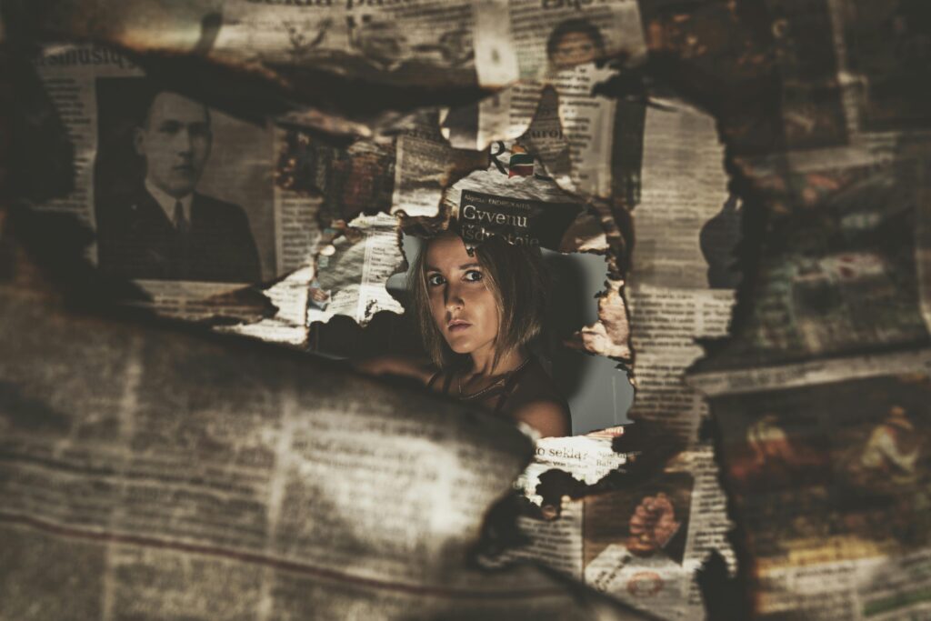 A woman's face peers through a torn hole in layered vintage newspaper clippings, her expression watchful and partially obscured by the aged paper surrounding her.