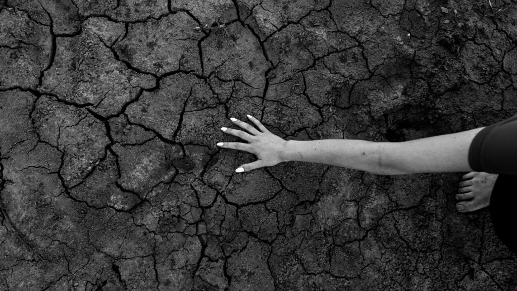Black and white photograph of an outstretched arm and hand reaching across deeply cracked, dry earth, conveying drought or desperation.
