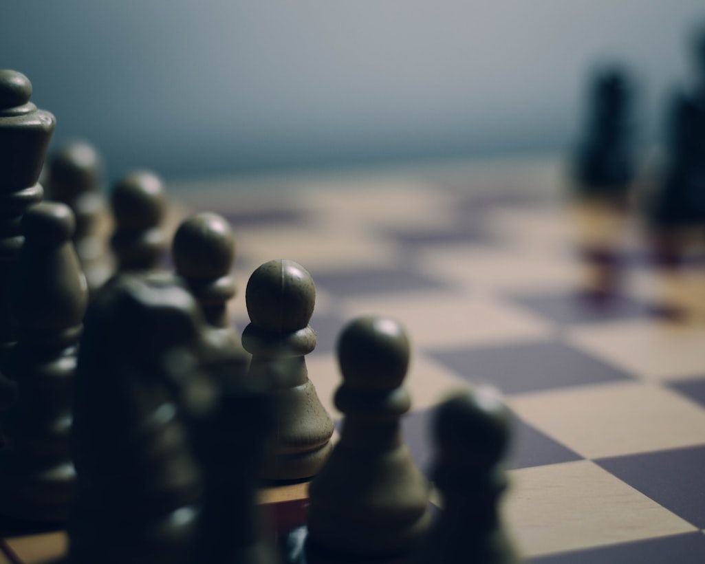 Chess pieces on a board photographed at eye level with shallow focus, pawns prominent in the foreground while other pieces blur into a moody grey background.