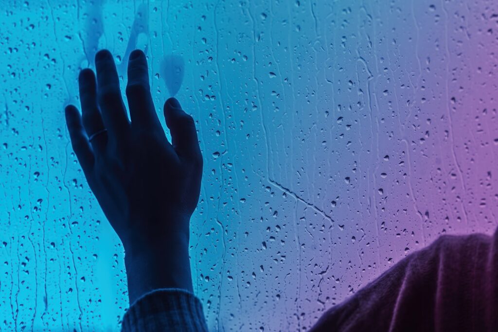 A hand pressed flat against a rain-streaked window, lit by blue and purple light that creates a melancholic, contemplative mood.