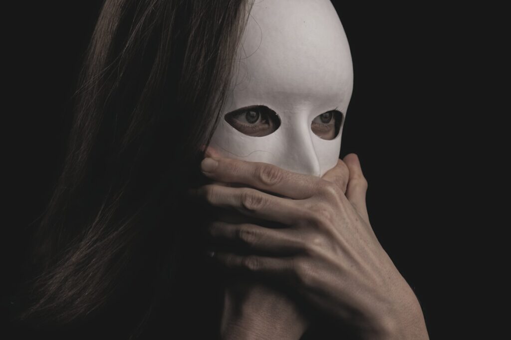A person with long dark hair holds a plain white theatrical mask over their face, only their eyes visible through the mask's eye holes against a black background.
