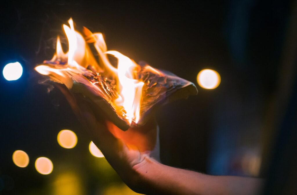 A hand holds burning paper or a photograph at night, flames consuming the material while soft bokeh lights glow in the dark background.