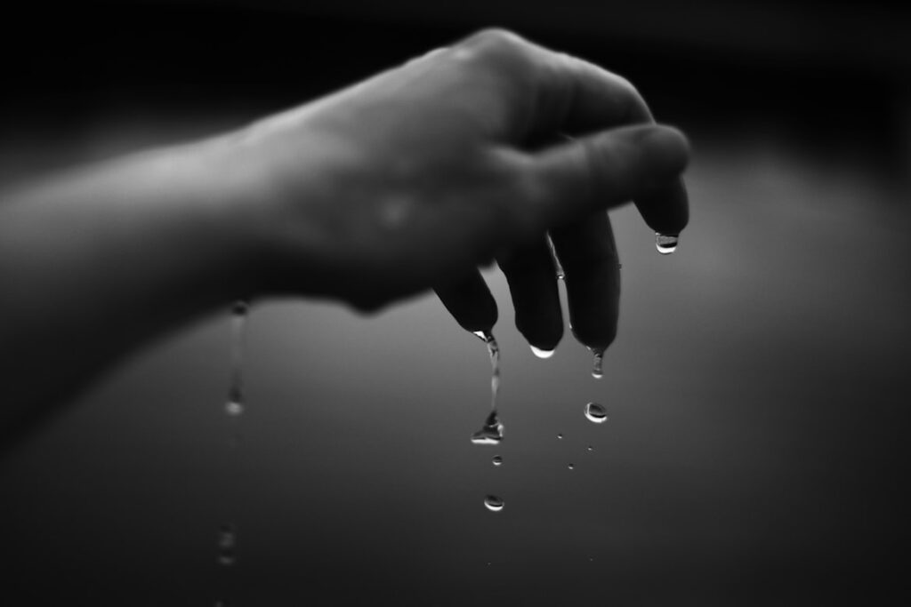 Black and white close-up of a hand with water droplets falling from the fingertips, each drop captured mid-descent against a soft grey background.