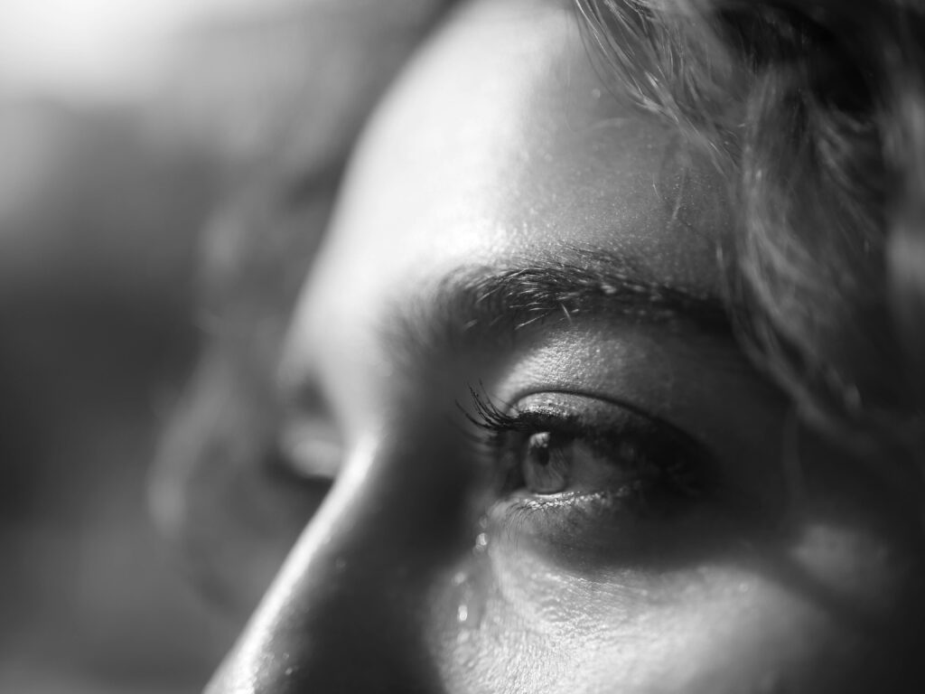 Black and white extreme close-up of a person's eye with a single tear rolling down their cheek, eyelashes and skin texture visible in intimate detail.