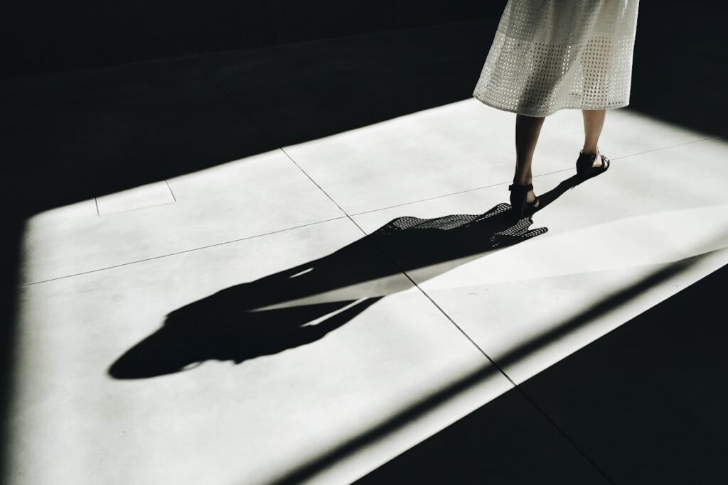 High contrast photograph of a person's lower legs and feet in a white skirt walking across a sunlit floor, their elongated shadow stretching dramatically beside them into darkness.