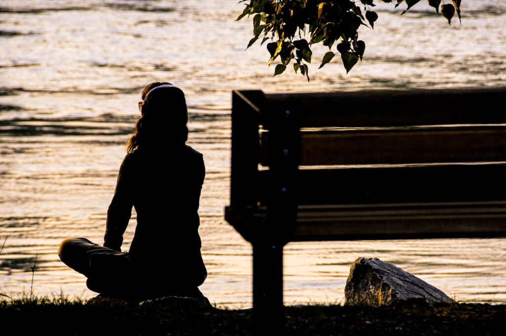 Silhouette of a woman sitting cross-legged on grass beside an empty bench, facing a body of water at sunset. Golden light reflects off the water's surface and catches the edges of her hair. Tree branches hang into the frame from above.