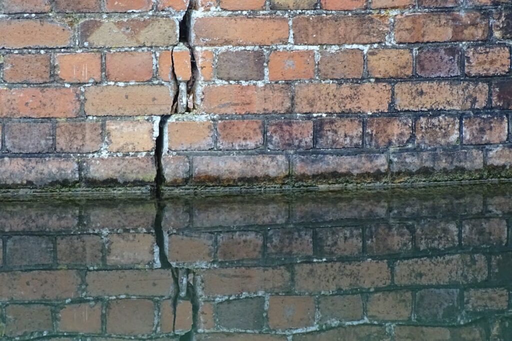 A weathered brick wall with a vertical crack running through its center, reflected in still water below. The bricks show patches of fading and discoloration, and moss grows along the waterline where the wall meets its own reflection.