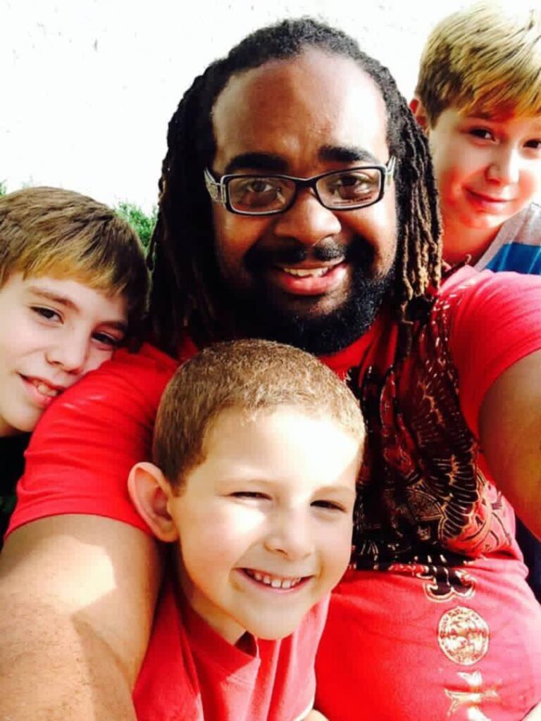 A man with dreadlocks and glasses takes a selfie with three smaller boys smiling.