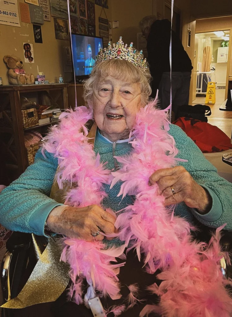 An elderly woman wearing a sparkly tiara and pink feather boa smiles joyfully in what appears to be a birthday celebration.
