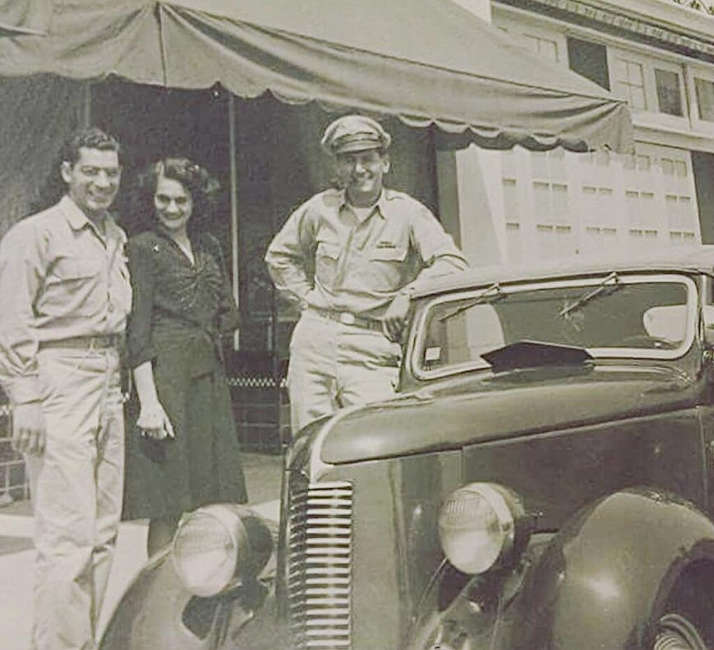 A vintage sepia photograph shows three people standing beside a 1940s-era car, with one man in military uniform.
