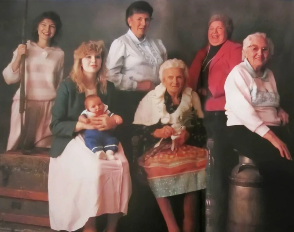 Six women spanning multiple generations pose for a family portrait, with the youngest holding a baby and two elderly women seated in front.
