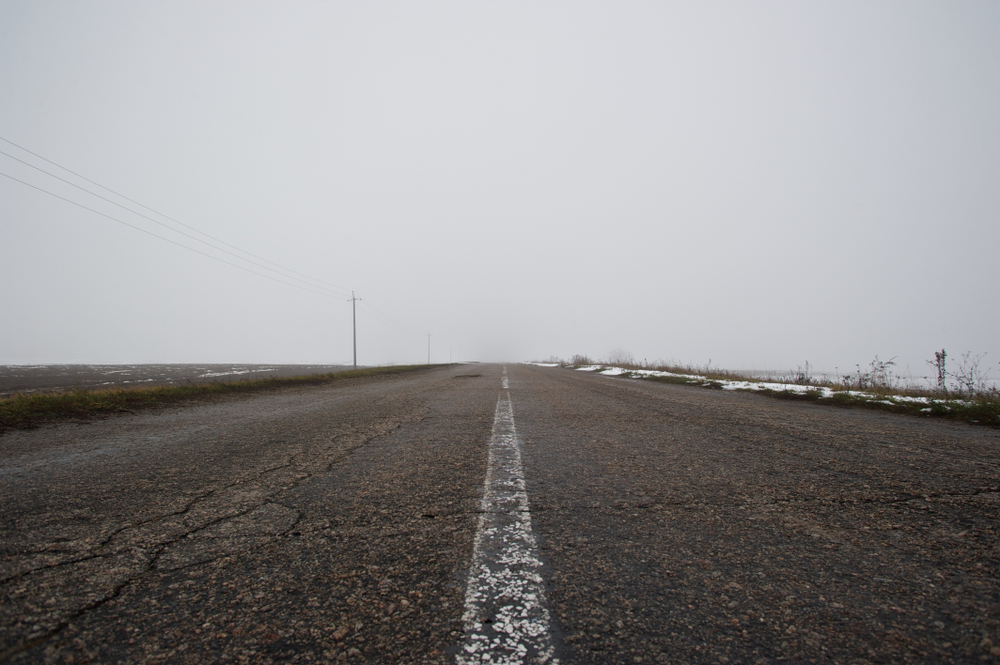 A picture of an empty road; cracks visible; faded road markings; grey sad sky in background.