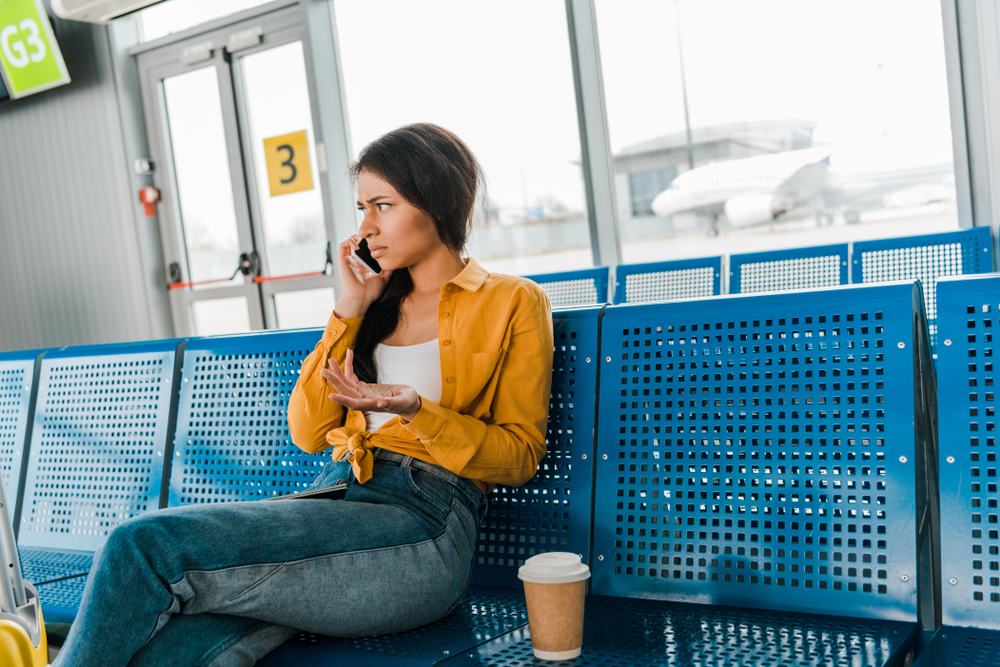 dissatisfied african american woman sitting in departure lounge with coffee to go and talking on smartphone