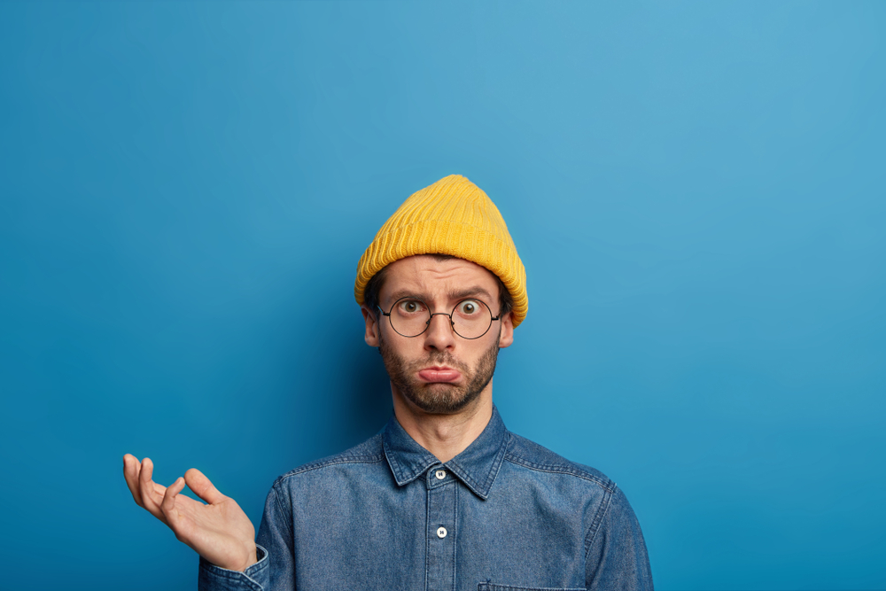Photo of upset puzzled man raises palm, looks with bothered sad expression, wears yellow hat, jean shirt, cannot agree with someones decision, isolated on blue background. Human reaction concept