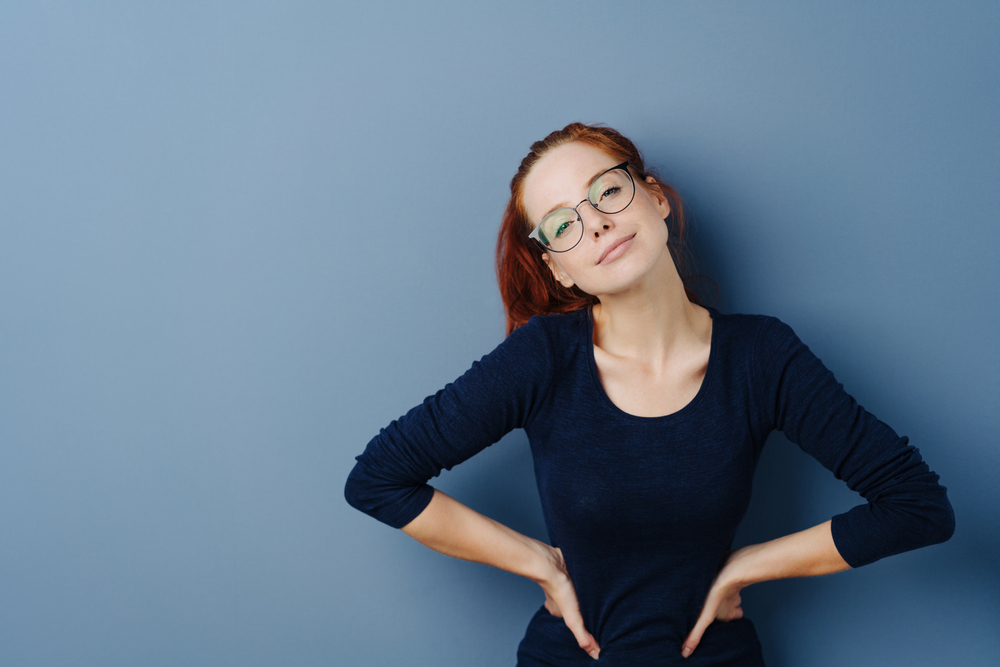 Pert self-assured young redhead woman wearing glasses standing with hands on hips looking at the camera with tilted head and quiet smile on a blue studio background with copy space