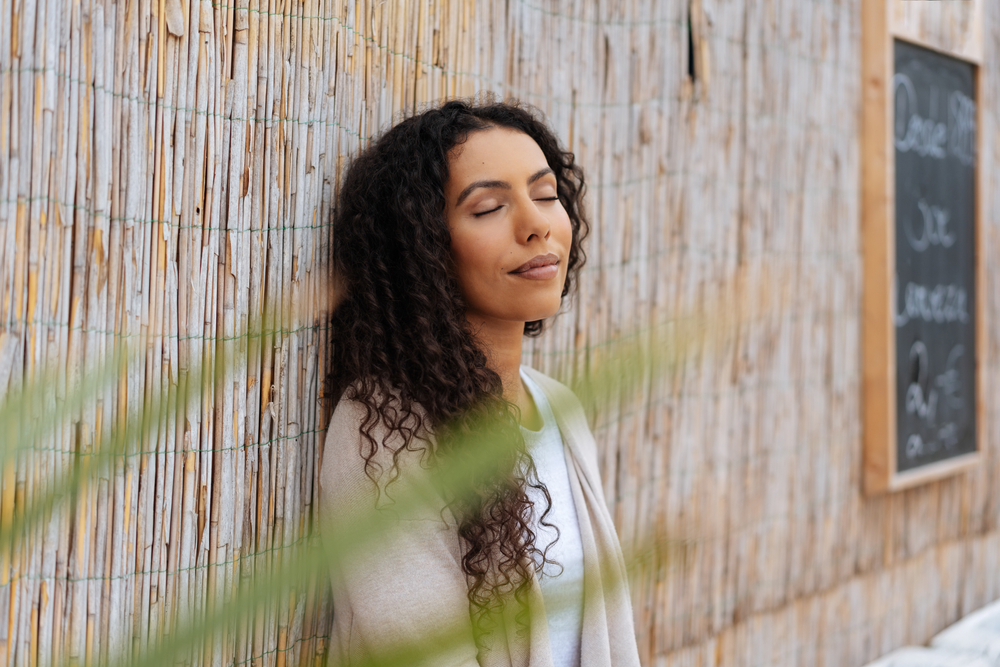 Young woman standing with closed eyes and a serene expression as she de-stresses releasing her inner tension against a feature reed wall in town