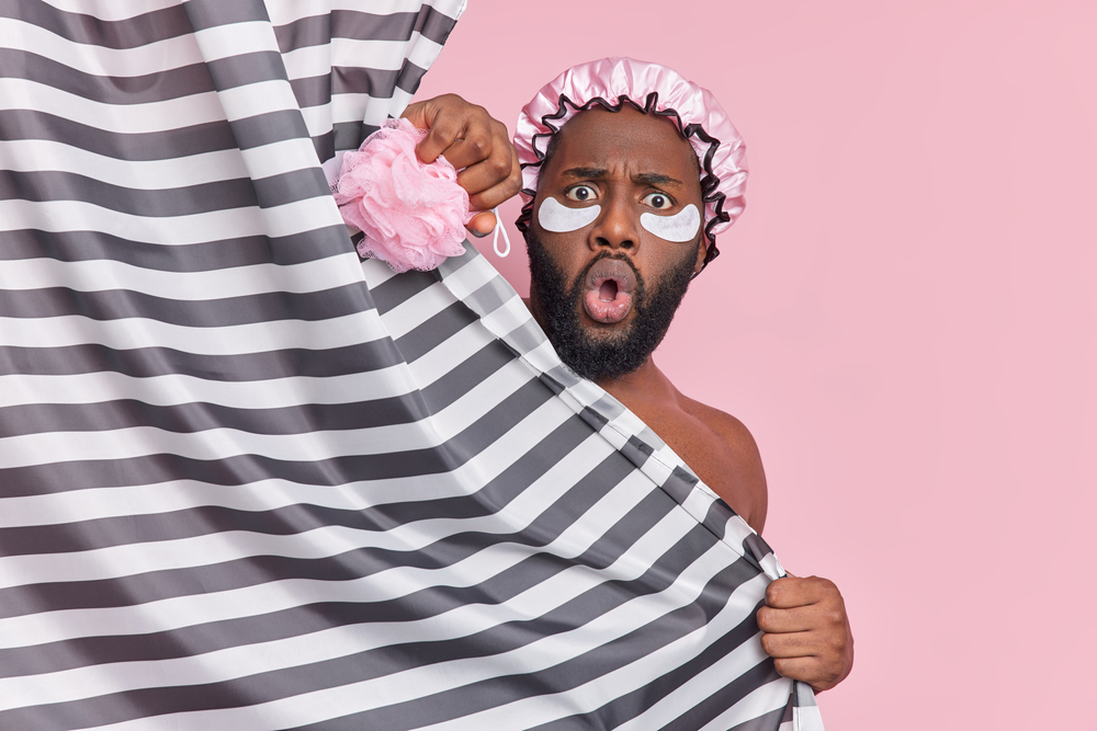 Shocked young Afro American man stunned someone came in bathroom hides naked behind striped shower curtain undergoes hygienic treatments applies beauty patches under eyes holds sponge says omg