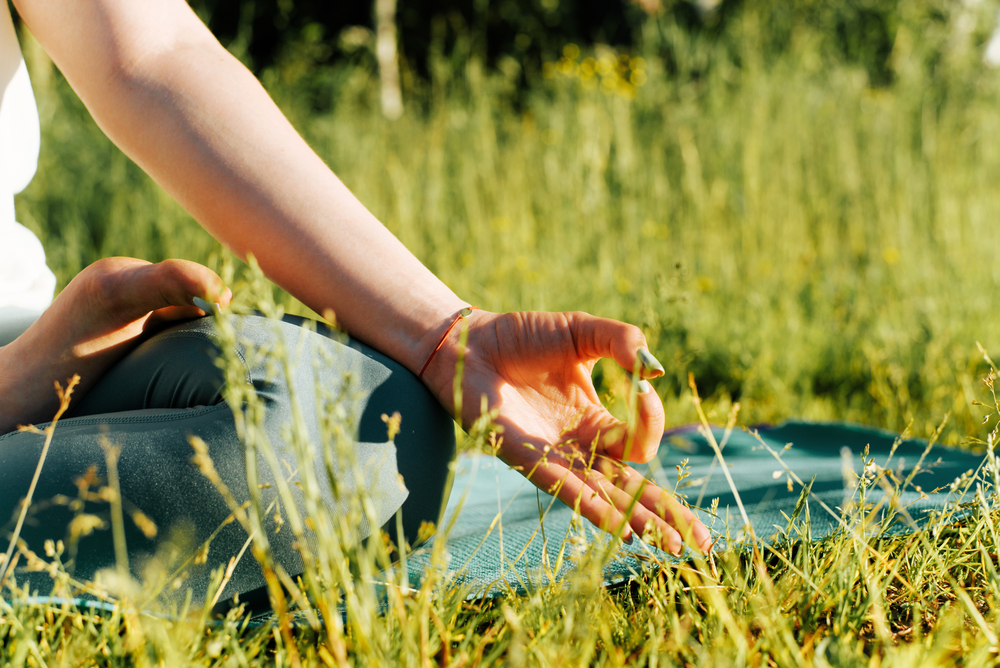 Meditation, outdoors. Close-up of a female hand in a yoga asana pose for Gyan Mudra meditation. Woman meditating in lotus position sitting on a rug on green grass in nature, cropped image.