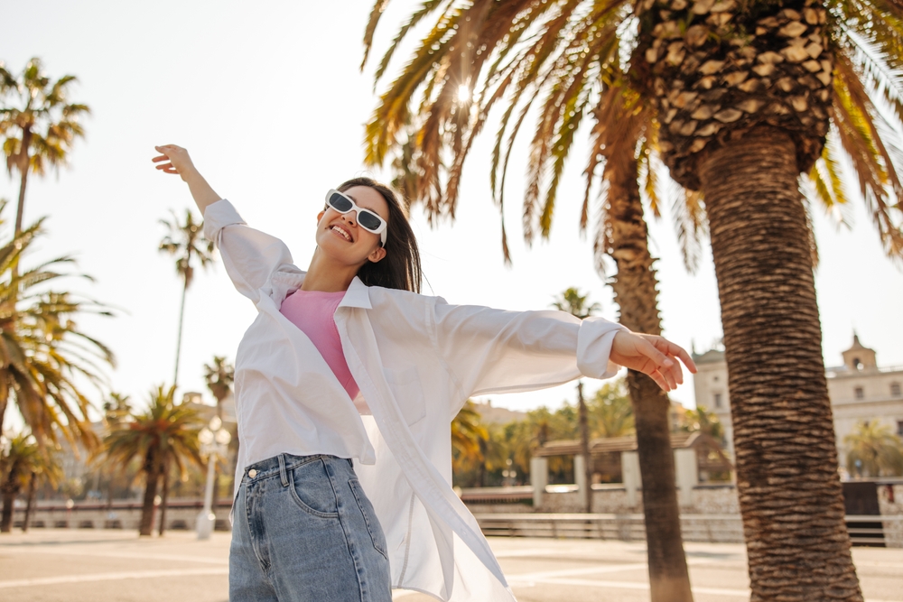 Joyful young caucasian woman waving her arms inhaling fresh air walking outdoors. Brunette wears sunglasses summer in sunny weather. Playful mood concept