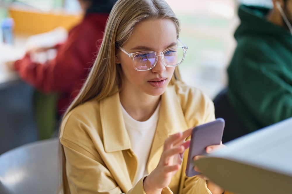 Teen girl gen z student using mobile phone looking at smartphone sitting at desk in university college campus classroom. Young blonde woman holding cellphone modern tech in university.
