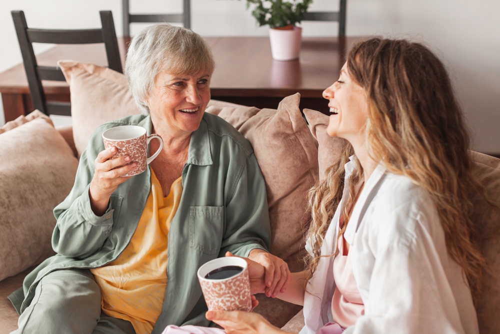 Smiling elderly mother and adult daughter sit on couch in living room drink hot tea coffee together, happy mature mom and grownup woman daughter relax on sofa at home, enjoy family weekend unity