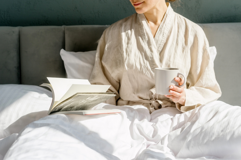 Woman seating on a bed, holding a cup of coffee, drinking espresso or herbal tea and reading book. Female in pajamas resting in hotel on weekend. Slow and simple living concepts. Cozy morning at home