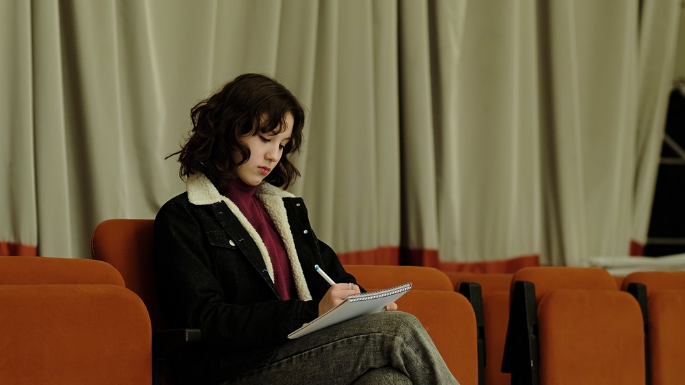 A young girl sits in a theater chair, focused on writing in a notebook. This image captures themes of creativity, concentration, and the pursuit of ideas in a calm environment.