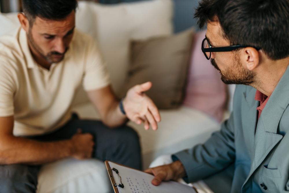 Psychologist with clipboard listening to his patient explaining his problems during therapy session on the sofa