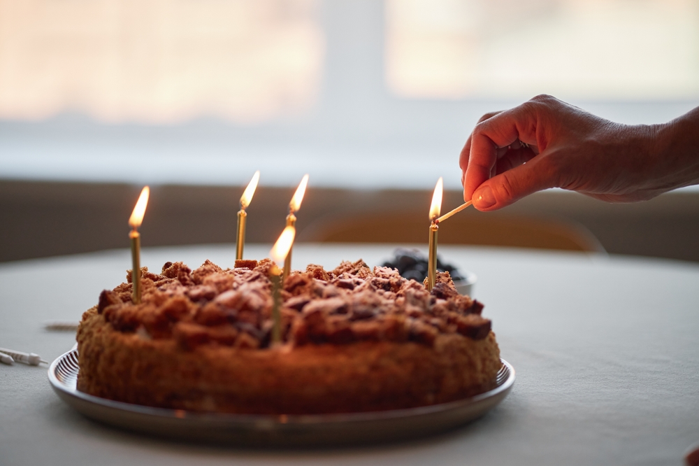 Caucasian adult hand lighting candles on birthday cake with matchstick, close up of fingers holding match above dessert, celebrating special occasion at home, blurred background visible