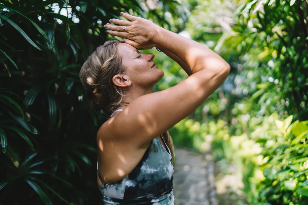 Woman standing in lush tropical path with hands to forehead and eyes closed, practicing focused gratitude and inner stillness through breath, light and nature-aligned awareness.