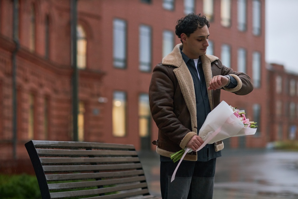 Young adult Caucasian man standing outdoors holding bouquet of flowers and checking wristwatch near bench in urban setting, appearing to wait for someone for dating meeting