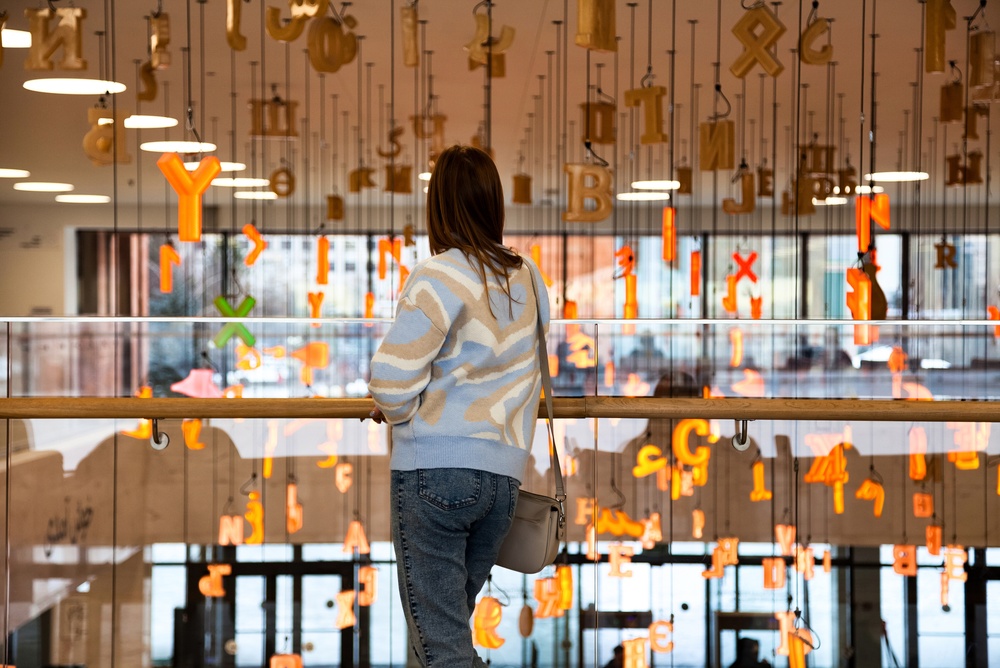 A young woman gazes upon floating letters and symbols at the Kazan National Library. She stands on a balcony, contemplating the alphabet art installation.