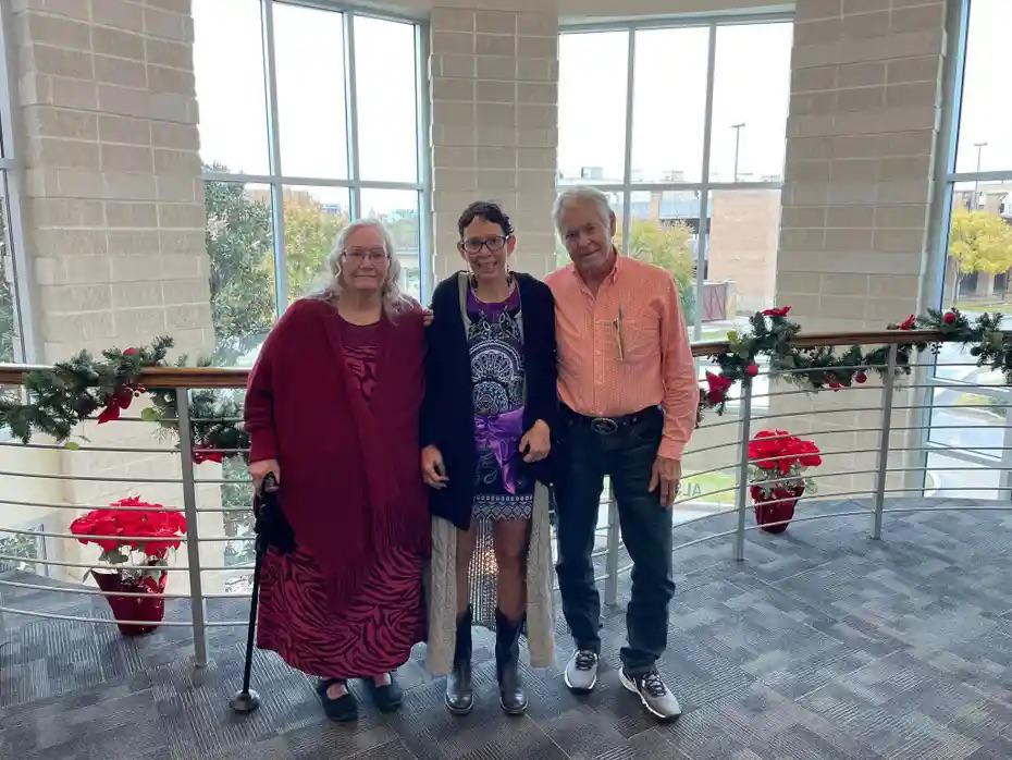 Three people stand together in a building lobby decorated with holiday garland. An older woman holds a cane on the left, a younger woman in a patterned dress stands in the middle, and an older man in an orange shirt is on the right.