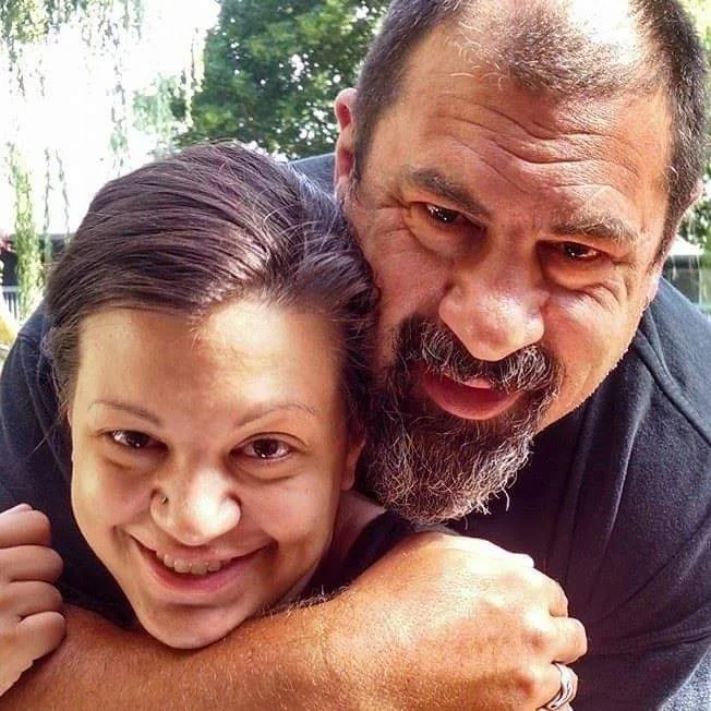 A close-up selfie of a couple smiling, the bearded man wrapping his arms around the woman from behind.
