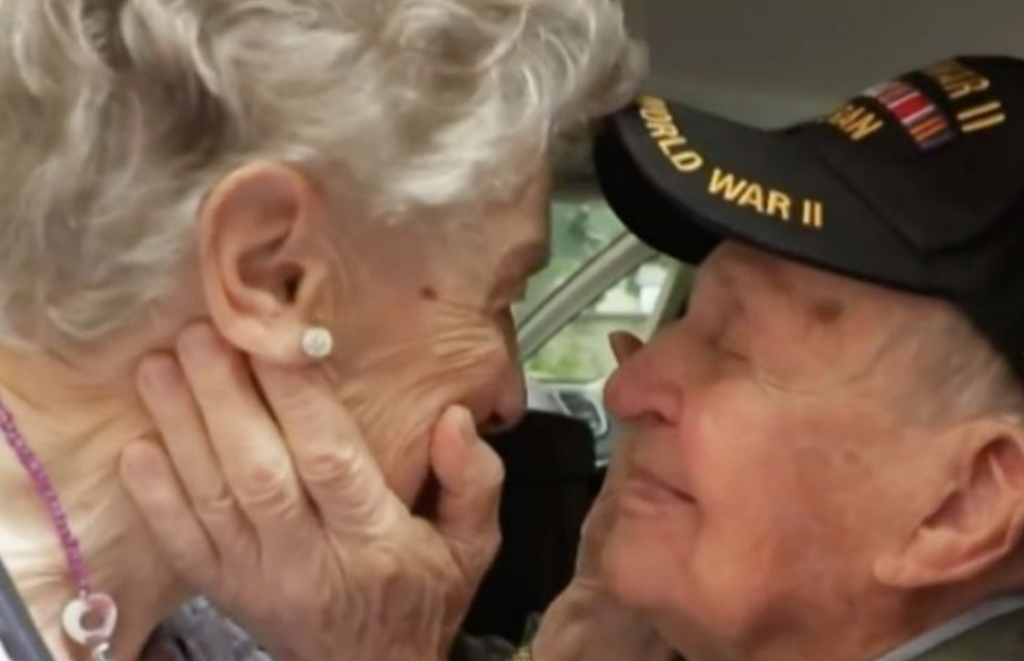 An elderly woman cups the face of a man wearing a World War II veteran cap, their foreheads nearly touching in a tender close-up.
