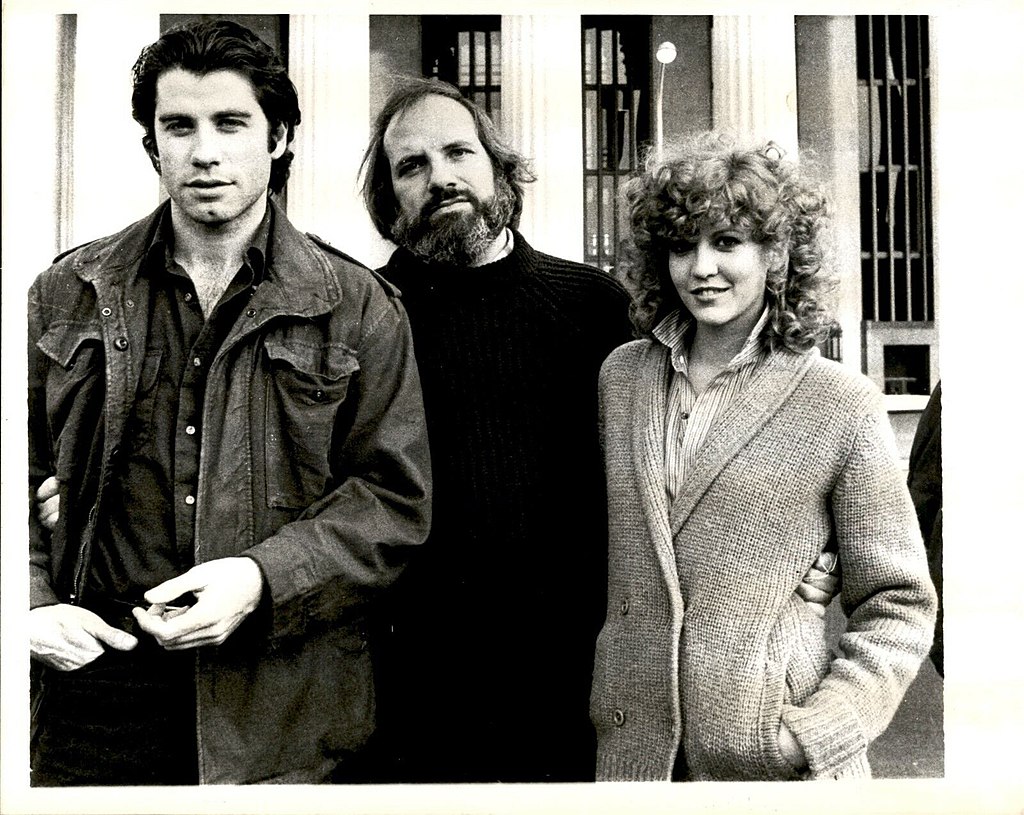 Black and white press photo of John Travolta with shaggy hair standing beside director Brian De Palma and actress Nancy Allen with curly blonde hair.