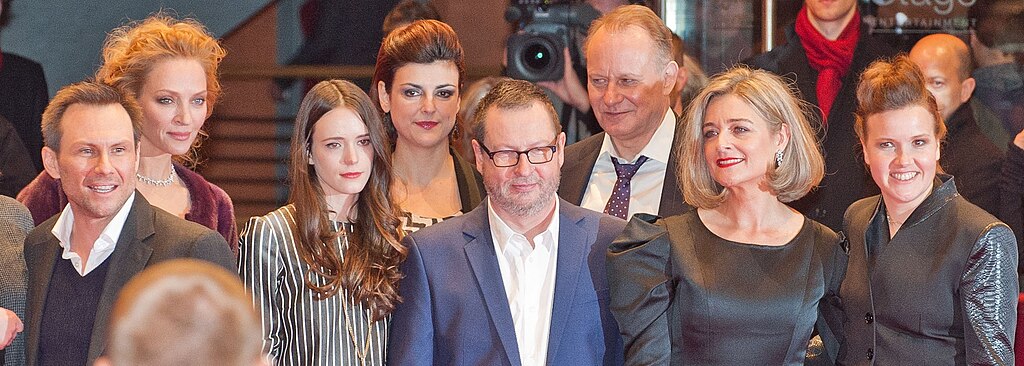 Director Lars von Trier stands in the center of a group photo with cast members at the Berlin premiere of Nymphomaniac in February 2014. Several actors in formal evening wear are arranged around him, with the warm lighting of the cinema lobby visible behind them.