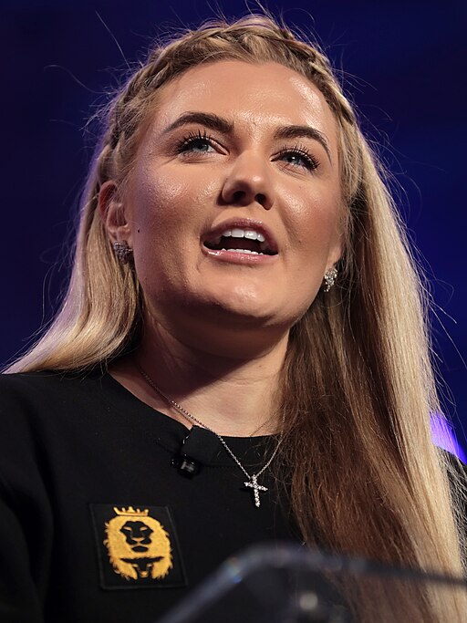 The Turning Point USA CEO addressing an audience from a podium, wearing a black top with a gold emblem and a cross necklace.