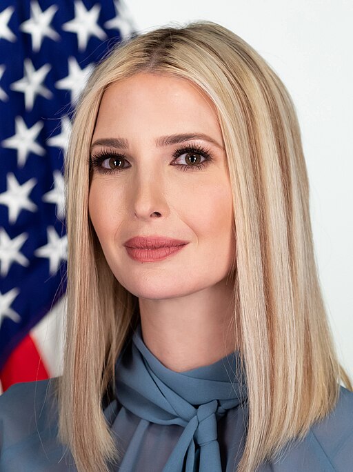 Ivanka Trump posing in front of an American flag, wearing a blue blouse with a bow tie collar.